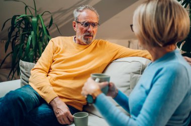Mutlu olgun karı koca, çay içerken oturma odasında rahat bir koltukta oturuyor. Yaşlı çift kanepede dinleniyor ve kahve içiyor. Modern gözlüklü sarı kazaklı adama odaklan.