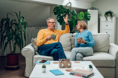 Mutlu olgun karı ve koca çay içip sohbet ederken oturma odasında rahat bir kanepede oturup dinleniyor. Yaşlı çift evdeki kanepede dinleniyor ve kahve içiyorlar. Boşluğu kopyala.