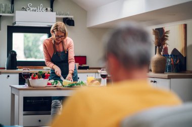 Yaşlı bir kadın, mutfakta sebze keserken, evde yemek pişirirken ve kocasıyla konuşurken ailesi için yemek hazırlıyor. Modern bir mutfakta son sınıf bir kadının portresi..