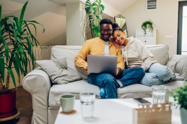 Genç, çok ırklı bir çift dizüstü bilgisayar kullanıyor ve haberleri tartışıyor ya da hafta sonları evdeki kanepede birlikte otururken online alışveriş yapıyor. Afrikalı Amerikalı erkek ve İspanyol kadın internette geziniyor..