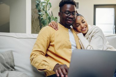 Laptop kullanan ve haberleri tartışan ya da hafta sonları evde online alışveriş yapan çok ırklı bir çiftin portresi. Afrikalı Amerikalı erkek ve İspanyol kadın internette geziniyor. Boşluğu kopyala.