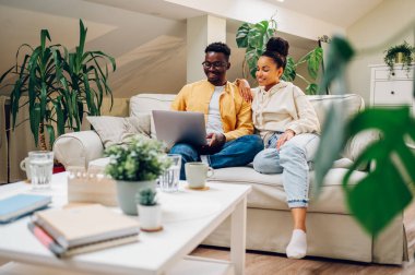 Farklı çiftler evlerinde kanepede yan yana otururken dizüstü bilgisayar kullanıyorlar. Gülümseyen Afro-Amerikan erkek ve İspanyol kadın hafta sonları birlikte vakit geçiriyorlar. Modern iç mimari.