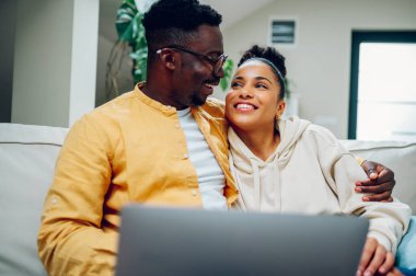 Evde rahatlayan ve dizüstü bilgisayar kullanan mutlu bir çiftin portresi. Gülümseyen Afrikalı Amerikalı koca ve İspanyol eş birlikte kanepede vakit geçirip birbirlerine bakıyorlar..