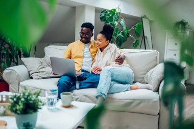Çeşitli çiftler evlerinde eğleniyor ve kanepede otururken dizüstü bilgisayar kullanıyorlar. Afrikalı Amerikalı erkek ve İspanyol kadın online alışveriş yapıyor ya da internette sörf yapıyor. Modern iç mekan. Boşluğu kopyala.