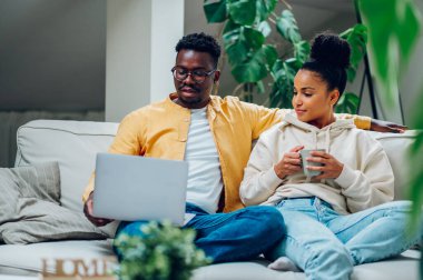 Genç, çok ırklı bir çift evdeki kanepede dizüstü bilgisayar kullanırken dinleniyor. Afrika kökenli Amerikalı erkek ve İspanyol kadın film izliyor ya da internette geziniyor ve kahve veya çay içiyor. Konforlu ev içi.