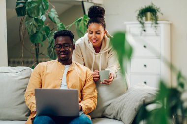 Genç, çok ırklı bir çift hafta sonu evde dizüstü bilgisayar ve internet gezintisi yapıyor. Kanepede oturan Afrikalı Amerikalı bir adam ve bir fincan kahve ya da çayla arkasında duran İspanyol bir kadın..