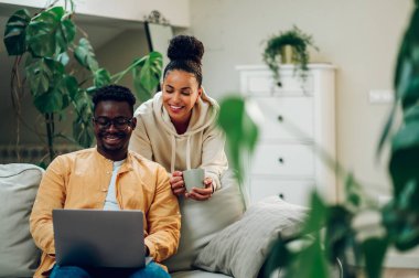 Evde vakit geçiren ve dizüstü bilgisayar kullanan farklı bir çiftin portresi. Afrikalı Amerikalı erkek ve İspanyol kadın online alışveriş yaparken ya da internette sörf yaparken birlikte eğleniyorlar. Boşluğu kopyala.