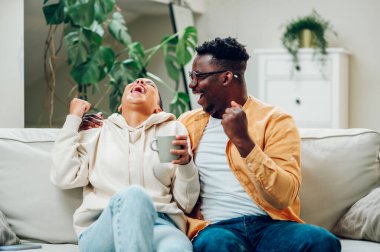 Mutlu İspanyol kadın ve afro-amerikan erkek evde televizyon izlerken tezahürat yapıyor ve gülüyorlar. Gülümseyen çok ırklı çift televizyonda spor izlemekten hoşlanıyor. Boşluğu kopyala.