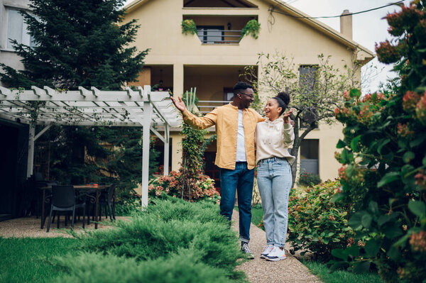 Happy diverse couple bought their new home and standing in front of it and showing keys. Copy space. Real estate owners. Hispanic woman and african american man posing in front of the house.
