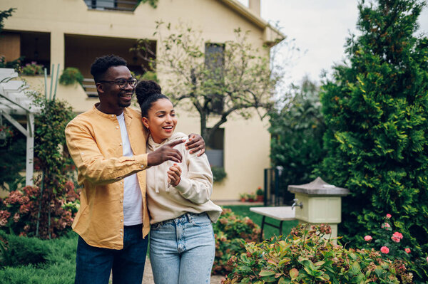 Overjoyed multiracial married couple standing in the garden in front of their new house. Happy african american husband embracing his hispanic wife and showing her neighborhood. Property ownership.