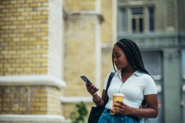 Güzel Afro-Amerikalı kadın akıllı telefon kullanıyor ve şehir dışındayken kahve içiyor.
