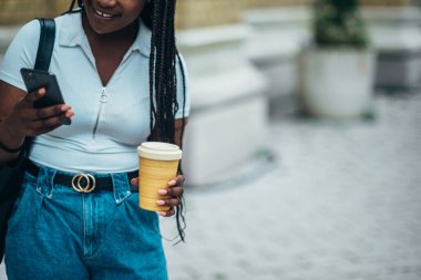 Neşeli Afro-Amerikalı kadın akıllı telefon kullanıyor ve şehir dışındayken kahve içiyor.