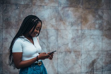 Neşeli Afrikalı Amerikalı kadın arka planda mermer bir duvarla şehirde akıllı telefon kullanıyor.