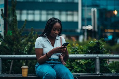 Neşeli Afro-Amerikalı kadın akıllı telefon kullanıyor ve şehir dışındayken kahve içiyor.