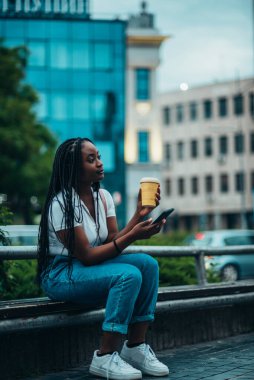 Güzel, Afro-Amerikalı bir kadın, akıllı telefon kullanıyor ve gündüzleri şehirde yürürken kahve içiyor.