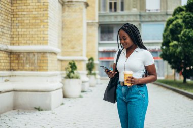 Güzel Afro-Amerikalı kadın akıllı telefon kullanıyor ve şehir dışındayken kahve içiyor.