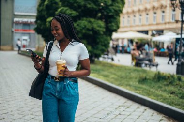 Güzel Afro-Amerikalı kadın akıllı telefon kullanıyor ve şehir dışındayken kahve içiyor.