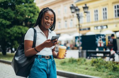 Neşeli Afro-Amerikalı kadın akıllı telefon kullanıyor ve şehir dışındayken kahve içiyor.