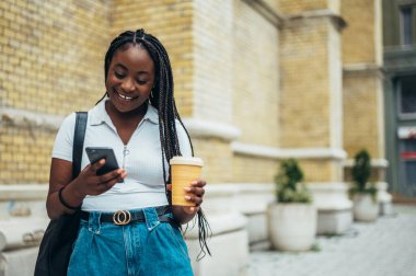 Neşeli Afro-Amerikalı kadın akıllı telefon kullanıyor ve şehir dışındayken kahve içiyor.