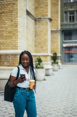 Neşeli Afro-Amerikalı kadın akıllı telefon kullanıyor ve şehir dışındayken kahve içiyor.