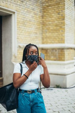 Genç, Afro-Amerikalı, koruyucu maske takan ve elinde akıllı bir telefon tutan bir kadın.