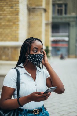 Güzel Afro-Amerikalı kadın koruyucu yüz maskesi takıyor ve salgın sırasında şehirde akıllı telefon kullanıyor.