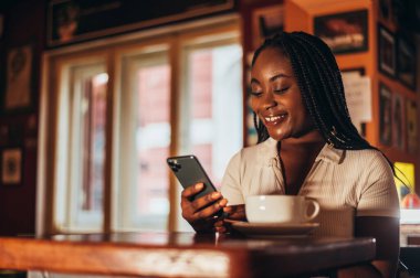 Bir kafede oturup kahve içerken akıllı telefon kullanan güzel, Afro-Amerikalı bir kadın.