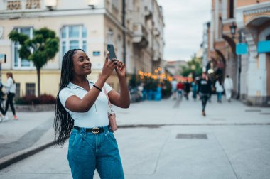 Neşeli Afrikalı Amerikalı kadın şehirde akıllı telefonuyla selfie çekiyor.