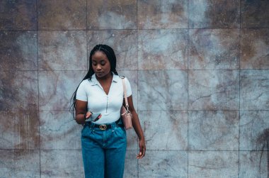 Neşeli Afrikalı Amerikalı kadın arka planda mermer bir duvarla şehirde akıllı telefon kullanıyor.