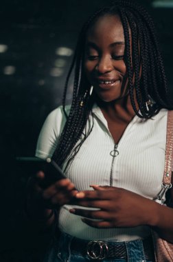 Metro istasyonunda toplu taşıma için beklerken akıllı telefon kullanan mutlu Afrikalı Amerikalı kadın.