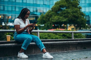 Neşeli Afro-Amerikalı kadın akıllı telefon kullanıyor ve şehir dışındayken kahve içiyor.