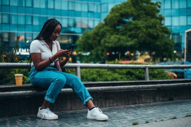 Neşeli Afro-Amerikalı kadın akıllı telefon kullanıyor ve şehir dışındayken kahve içiyor.