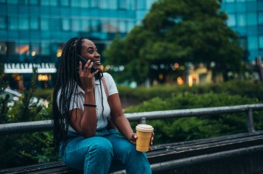 Güzel, Afro-Amerikalı bir kadın şehirde bir bankta otururken akıllı telefon kullanıyor ve kahve içiyor.