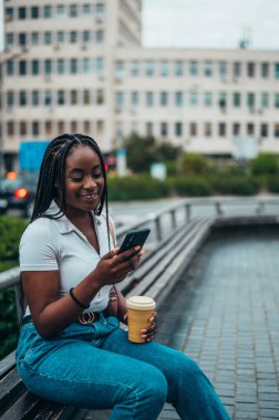 Neşeli Afro-Amerikalı kadın şehir dışındayken akıllı telefon ve kahve servisi kullanıyor.