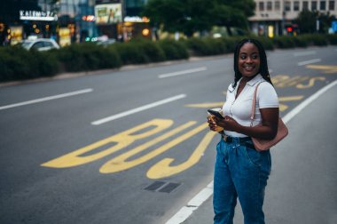 Güzel Afrikalı Amerikalı kadın otobüs durağında dikilip şehirde toplu taşıma için beklerken akıllı telefon kullanıyor.