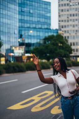 Neşeli Afro-Amerikalı kadın akıllı telefon kullanıyor ve şehirde yol kenarında taksi çağırıyor.