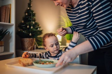 Sevimli küçük bir çocuk evde yemek masasında oturuyor ve annesi ona yardım ederken yemeğini yiyor. Aç çocuk sağlıklı yemeğini evde, yemek masasında yerken annesi ona yardım ediyor..