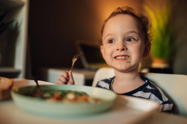Yemek masasında elinde çatalla oturan ve yemeğini yerken gülümseyen sevimli bir çocuğun portresi. Mutlu bir çocuğun evde sağlıklı bir öğle yemeği yemesine yakın..