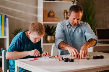 Oğlu ve babasının boş zamanlarını evde geçirdikleri geniş bir görüntü. Küçük oyuncak parçalarını sabırla bir araya getiren genç bir çocuğa odaklan. Hafta sonunu evde birlikte geçirmek. Aile kavramı.
