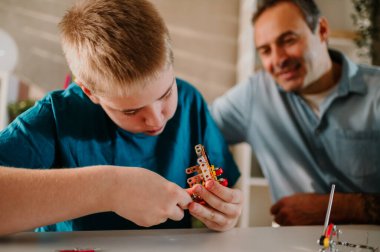Sarışın bir gencin oyuncağını bitirirken yakın plan çekimleri. Son parçaları yerleştirirken oyuncağı tutan parmaklarına odaklan. Babasının mutlulukla izlediği bulanık arka planı.