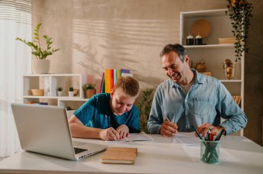 Mutlu babasıyla yemek masasında oturan gülümseyen genç bir sarışının ev ödevini yaparken ve evdeki güneşli günün keyfini çıkarırken geniş bir açıyla. İkisi de aşağı bakıyor ve gülüyor. Boşluğu kopyala.