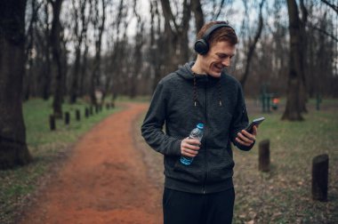 Genç kızıl saçlı adam doğada koşarken akıllı telefon ve kulaklık kullanıyor. Spor ve spor konsepti, sağlıklı yaşam tarzı, motivasyon..