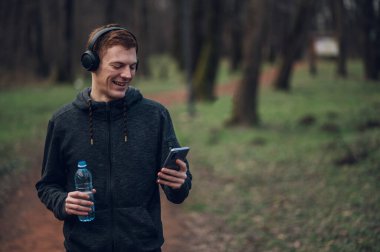 Genç kızıl saçlı adam doğada koşarken akıllı telefon ve kulaklık kullanıyor. Spor ve spor konsepti, sağlıklı yaşam tarzı, motivasyon..