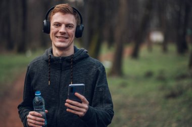 Atletik kızıl saçlı genç adam orman yolunda koşarken akıllı telefon kullanıyor. Koşarken kulaklıkla müzik dinlemek. Atletik ve sağlıklı. Kameraya bakıyorum..