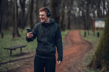 Genç kızıl saçlı adam doğada koşarken akıllı telefon ve kulaklık kullanıyor. Spor ve spor konsepti, sağlıklı yaşam tarzı, motivasyon..