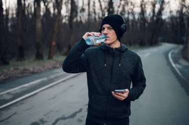 Kızıl saçlı atlet antrenmanına ara verip su içiyor ve ormanda çalışırken akıllı telefon kullanıyor. Sağlıklı yaşam tarzı kavramı.
