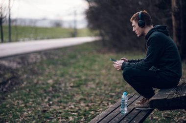 Atletik kızıl saçlı genç adam ormanda koşarken akıllı telefon kullanıyor. Koşarken kulaklıkla müzik dinlemek. Atletik ve sağlıklı..