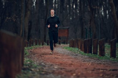 Kızıl saçlı erkek, doğa harikası bir orman yolunda maraton koşuyor ve antrenman yapıyor. Aktif spor konsepti. Dışarıdaki eğitiminde kulaklık kullanıp müzik dinleyen bir koşucu..