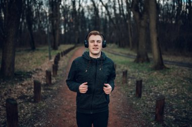 Kızıl saçlı erkek, doğa harikası bir orman yolunda maraton koşuyor ve antrenman yapıyor. Aktif spor konsepti. Dışarıdaki eğitiminde kulaklık kullanıp müzik dinleyen bir koşucu..