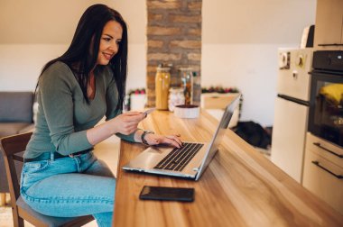 Mutfakta otururken dizüstü bilgisayar ve kredi kartı kullanan gülümseyen orta yaşlı bir kadın. İnternet üzerinden alışveriş yaparken ödeme sistemi üzerinden bilgi girişi. İnternetten alışveriş.